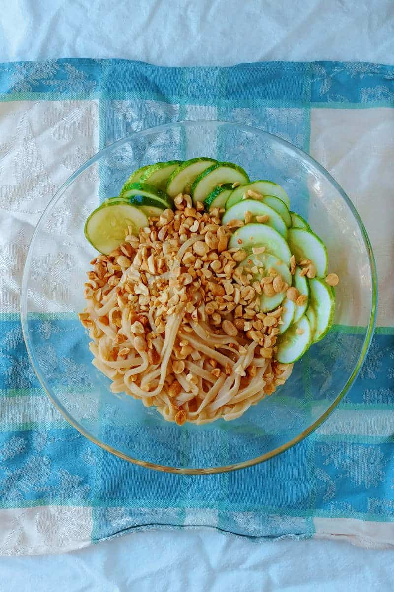 sesame noodles, topped with peanuts and cucumbers, in a glass bowl on a napkin