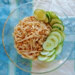 sesame noodles, topped with peanuts and cucumbers, in a glass bowl