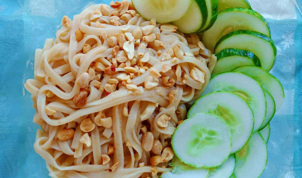 sesame noodles, topped with peanuts and cucumbers, in a bowl, close-up