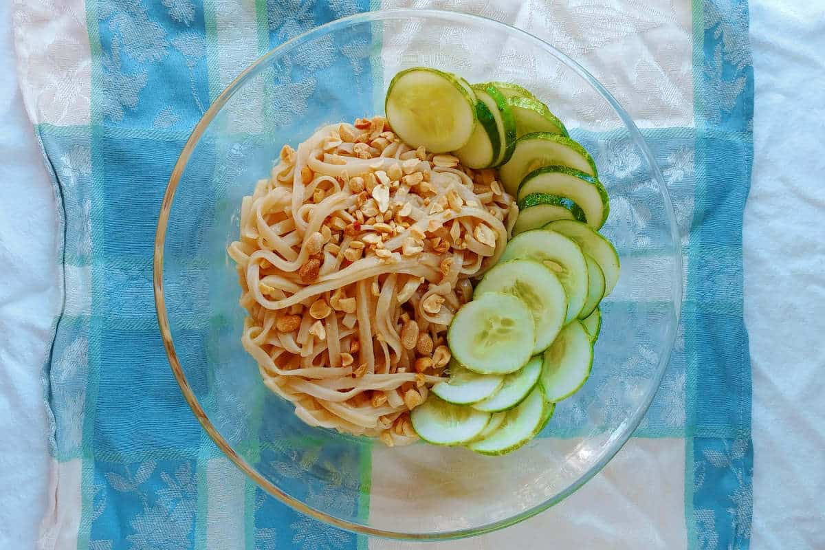 sesame noodles, topped with peanuts and cucumbers, in a glass bowl