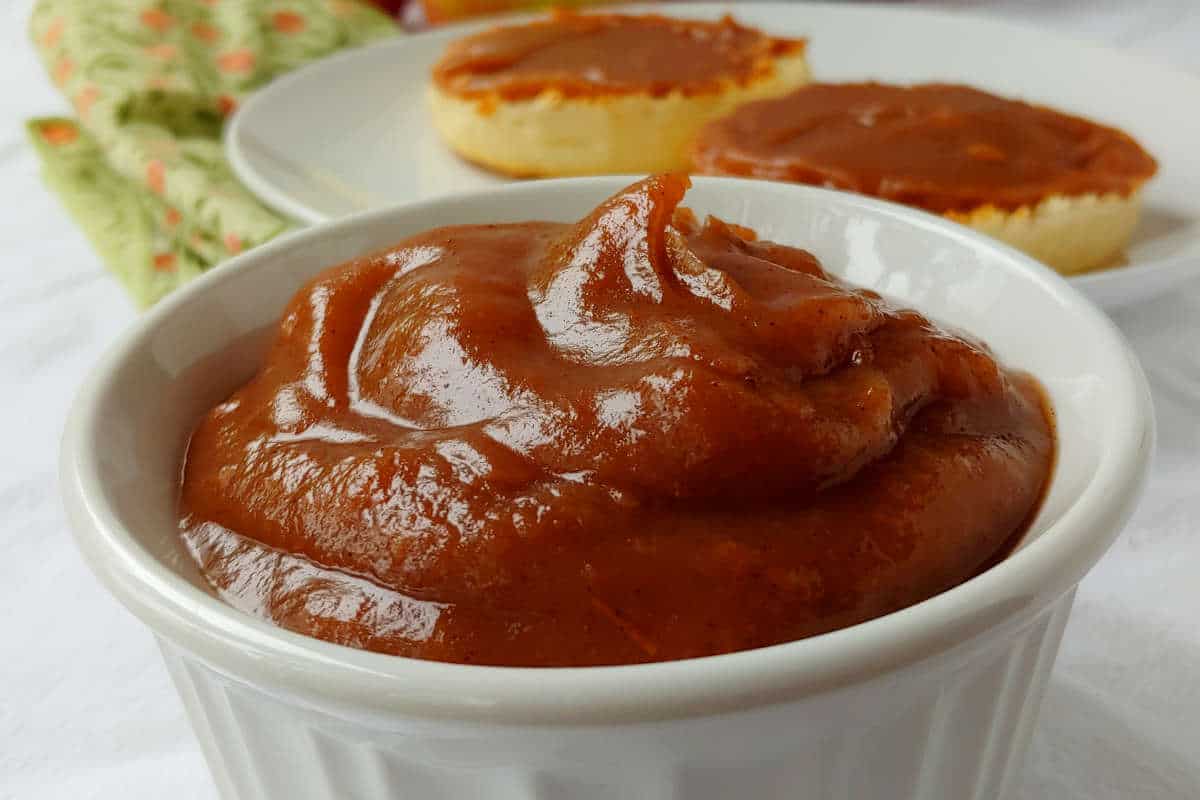 no-sugar-added apple butter, in a white bowl, with english muffins spread with apple butter in the background