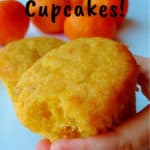 a hand holding a vegan orange cupcake with a bite taken from it, with oranges in the background - pin for Pinterest