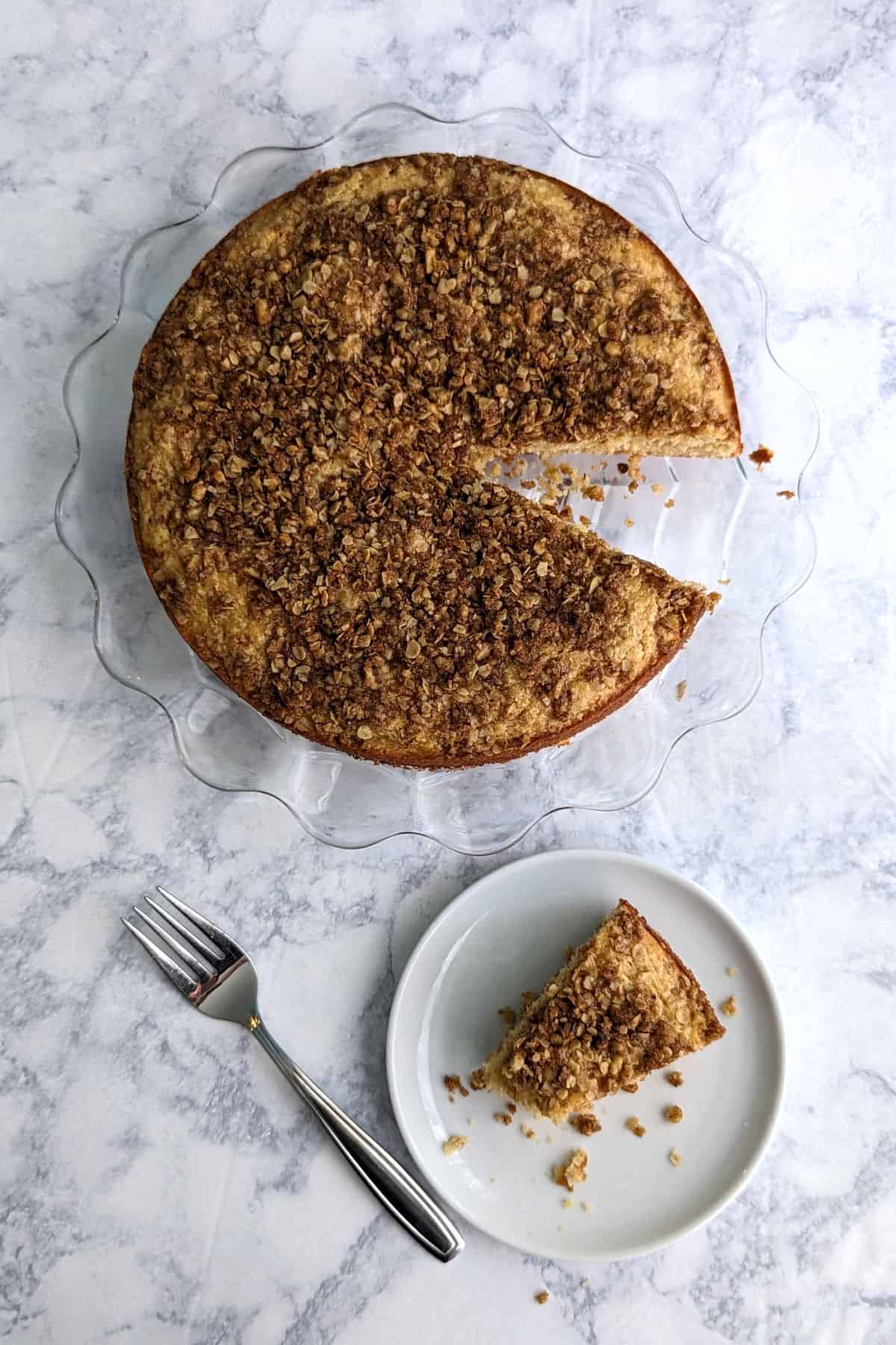 slice of sour cream coffee cake with a bite missing on a serving plate, with fork next to it. Whole cake is missing a slice.