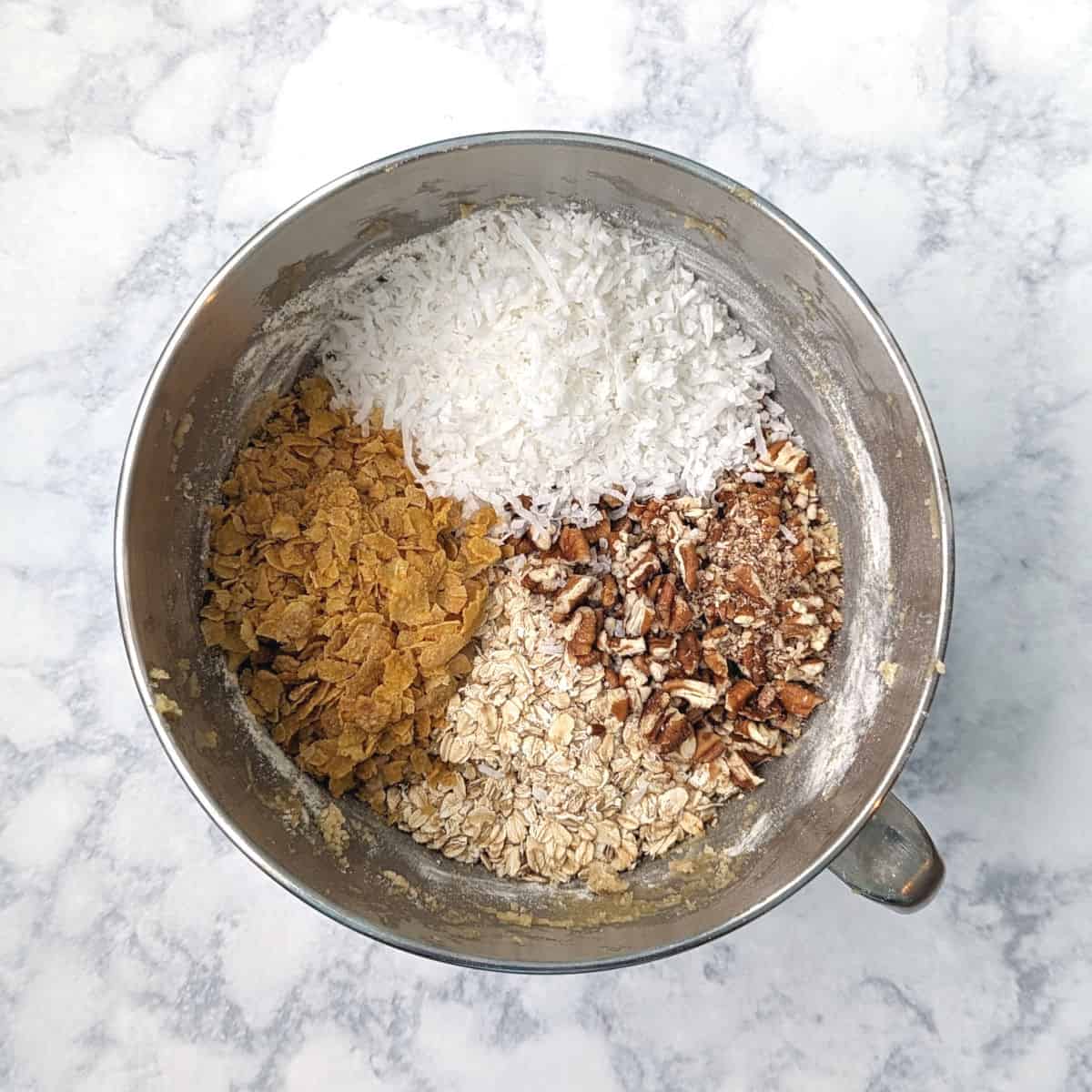 oats, pecans, coconut, and corn flakes, on top of cookie dough in a stand mixer bowl