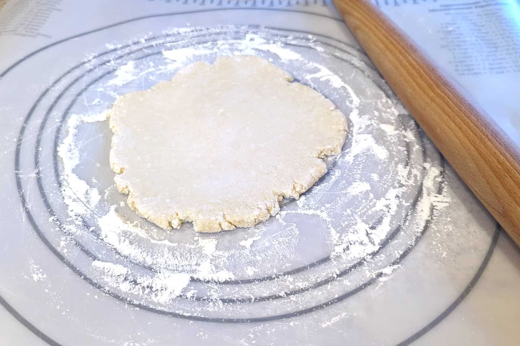 disc of pie crust dough, having been rolled out by about 1 inch, on a rolling mat with a rolling pin along the side