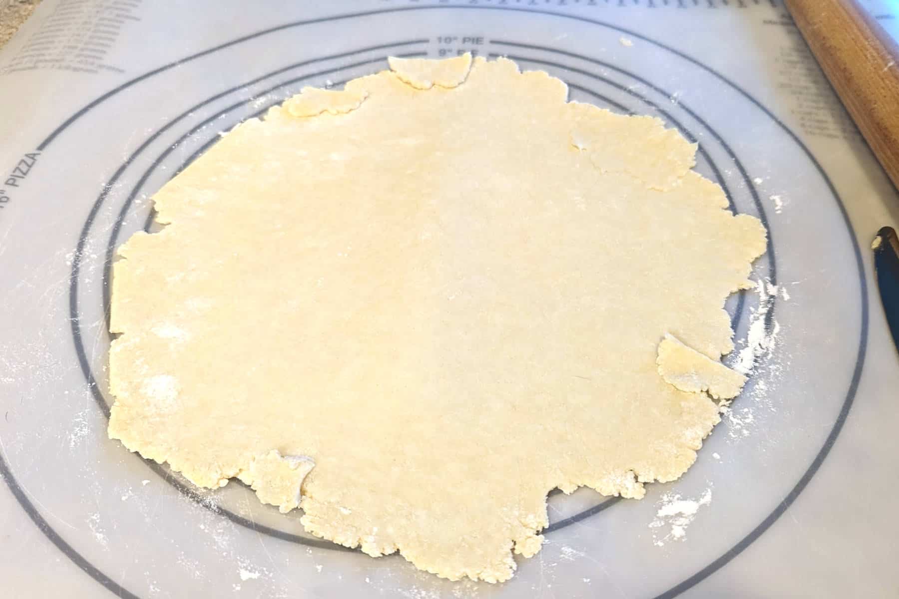 pie crust dough almost rolled out all the way. Uneven pieces of dough have been cut off of the edges and used as patches where there are gaps