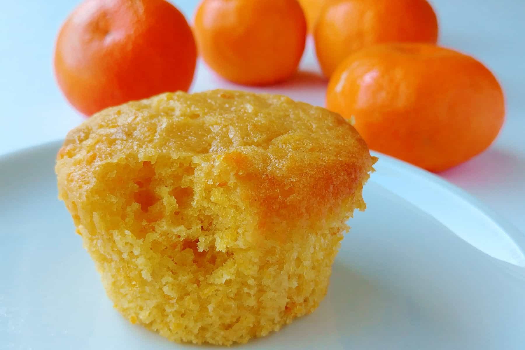 A vegan orange muffin with a small bite out of it, on a white place. Behind it are additional clementines.