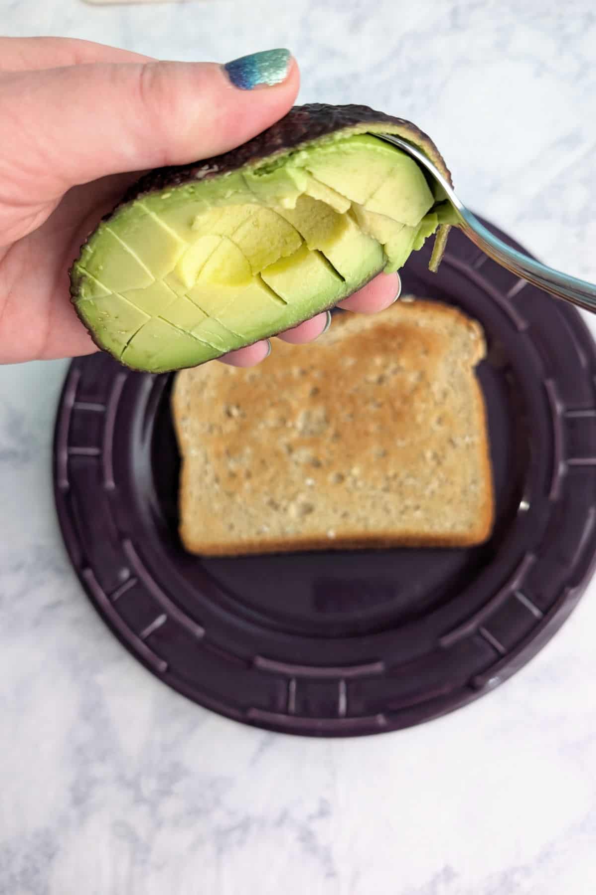 A spoon scoops cubed avocado from the skin, to pile onto the slice of toast below.