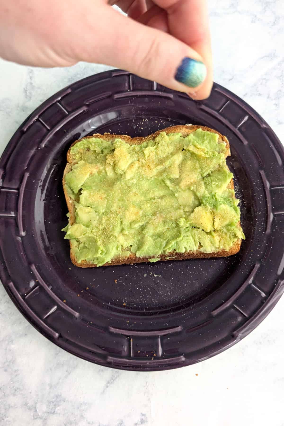 Fingers sprinkle salt onto a piece of avocado toast that already has garlic powder on it.