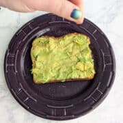 A hand sprinkles salt and garlic powder onto a slice of avocado toast.