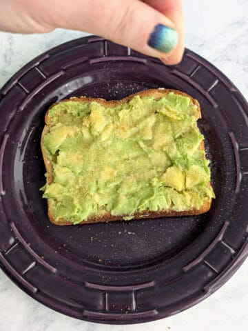 A hand sprinkles salt and garlic powder onto a slice of avocado toast.