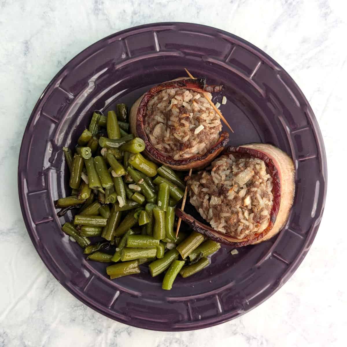 Two Fake Steak patties, wrapped in bacon, alongside green beans, on a purple plate.