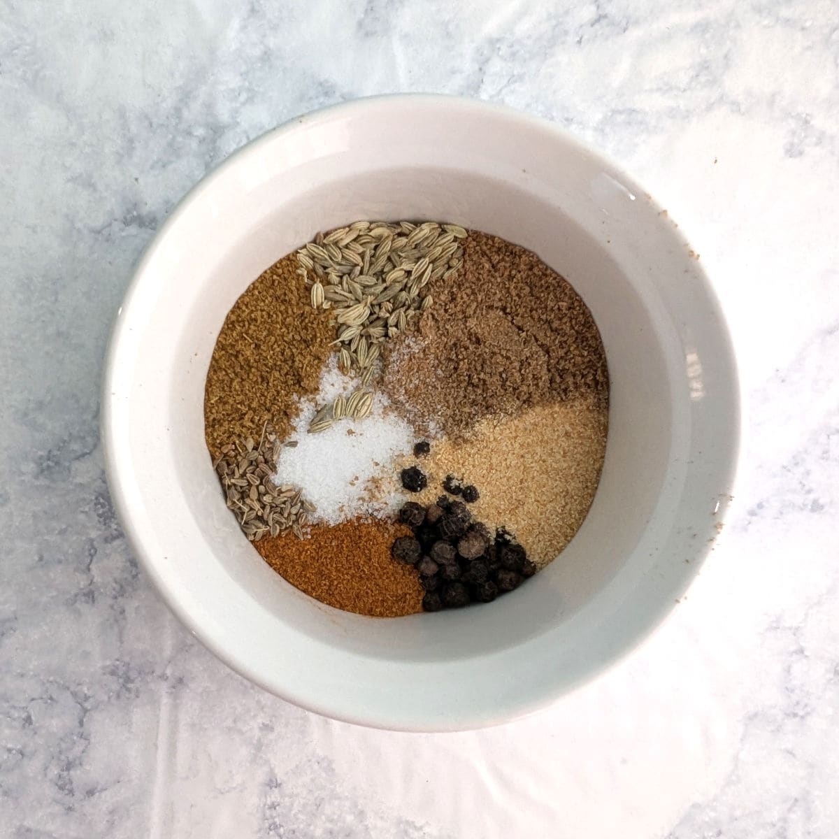 Fennel seeds, ground cumin, ground coriander, garlic powder, anise seeds, whole black pepper, cayenne, and salt, in a small white ramekin.