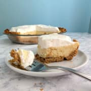 A fork with a bite of key lime pie rests on the plate with the remaining slice. The rest of the pie is in the background.