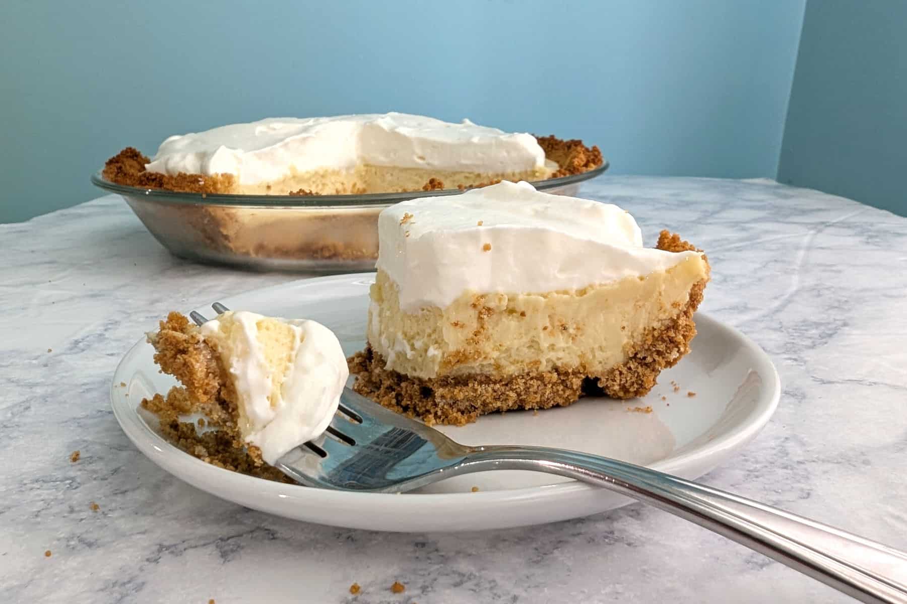 A fork with a bite of key lime pie rests on the plate with the remaining slice. The rest of the pie is in the background.