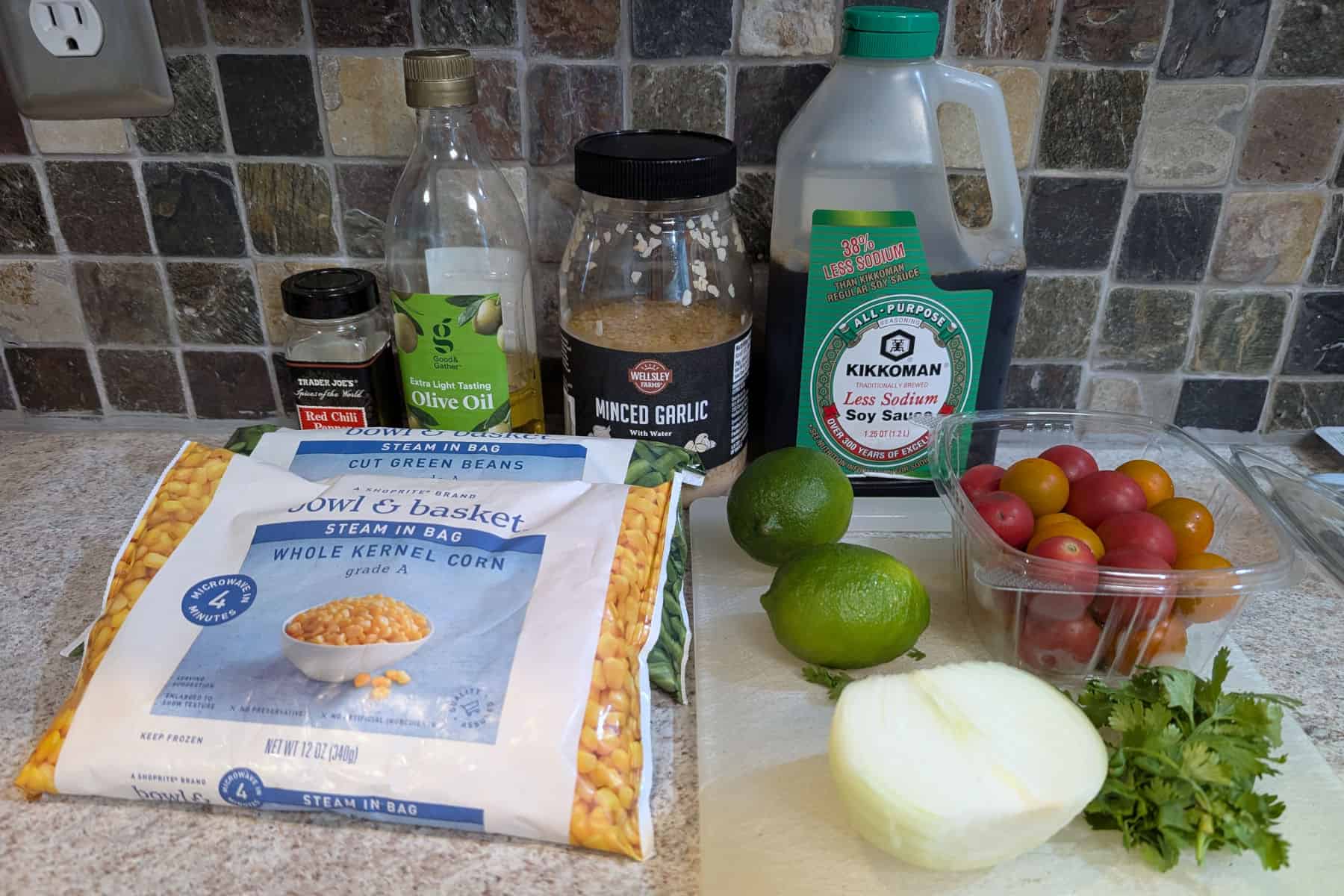 Ingredients for Corn and Green Bean Salad with Lime: frozen corn, frozen green beans, red pepper flakes, olive oil, minced garlic, soy sauce, limes, onion, tomatoes, and cilantro.