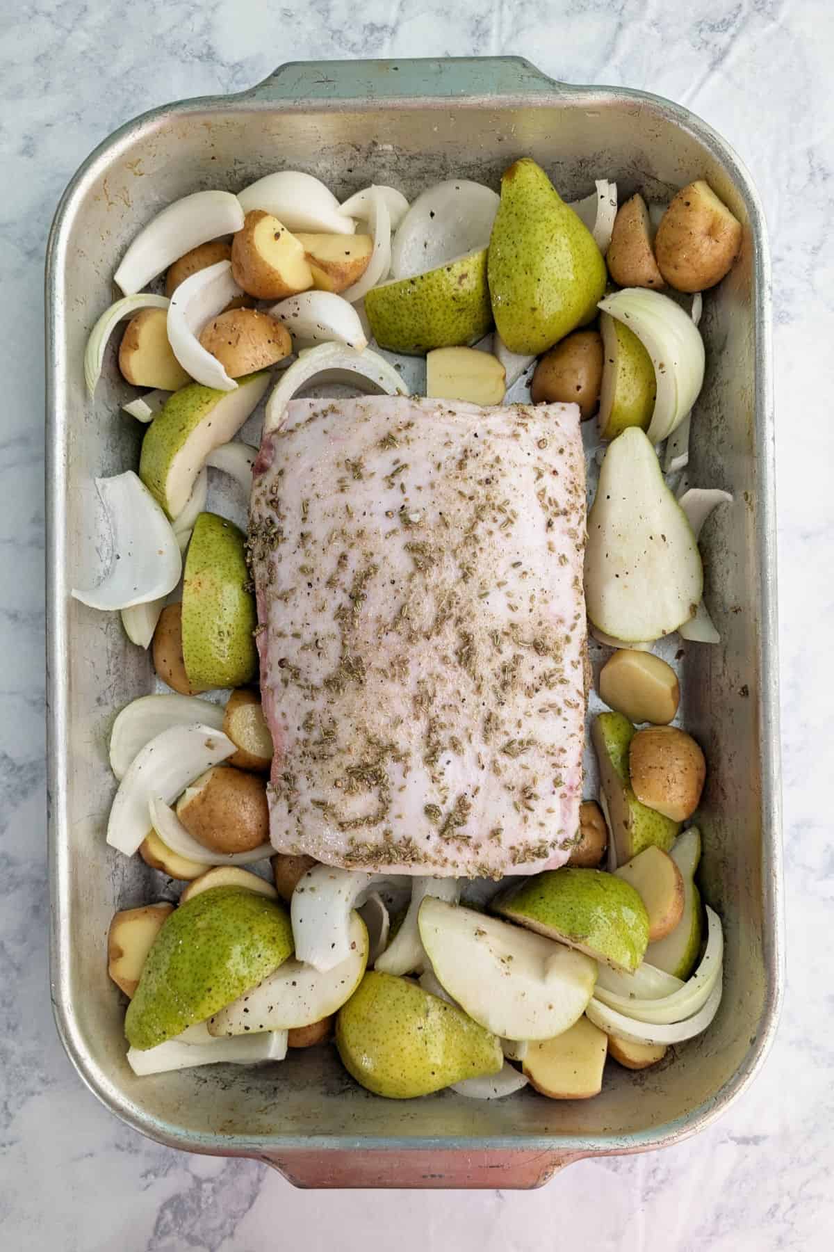 A raw pork loin surrounded by halved potatoes, quartered pears, and sliced onions, in a roasting pan.