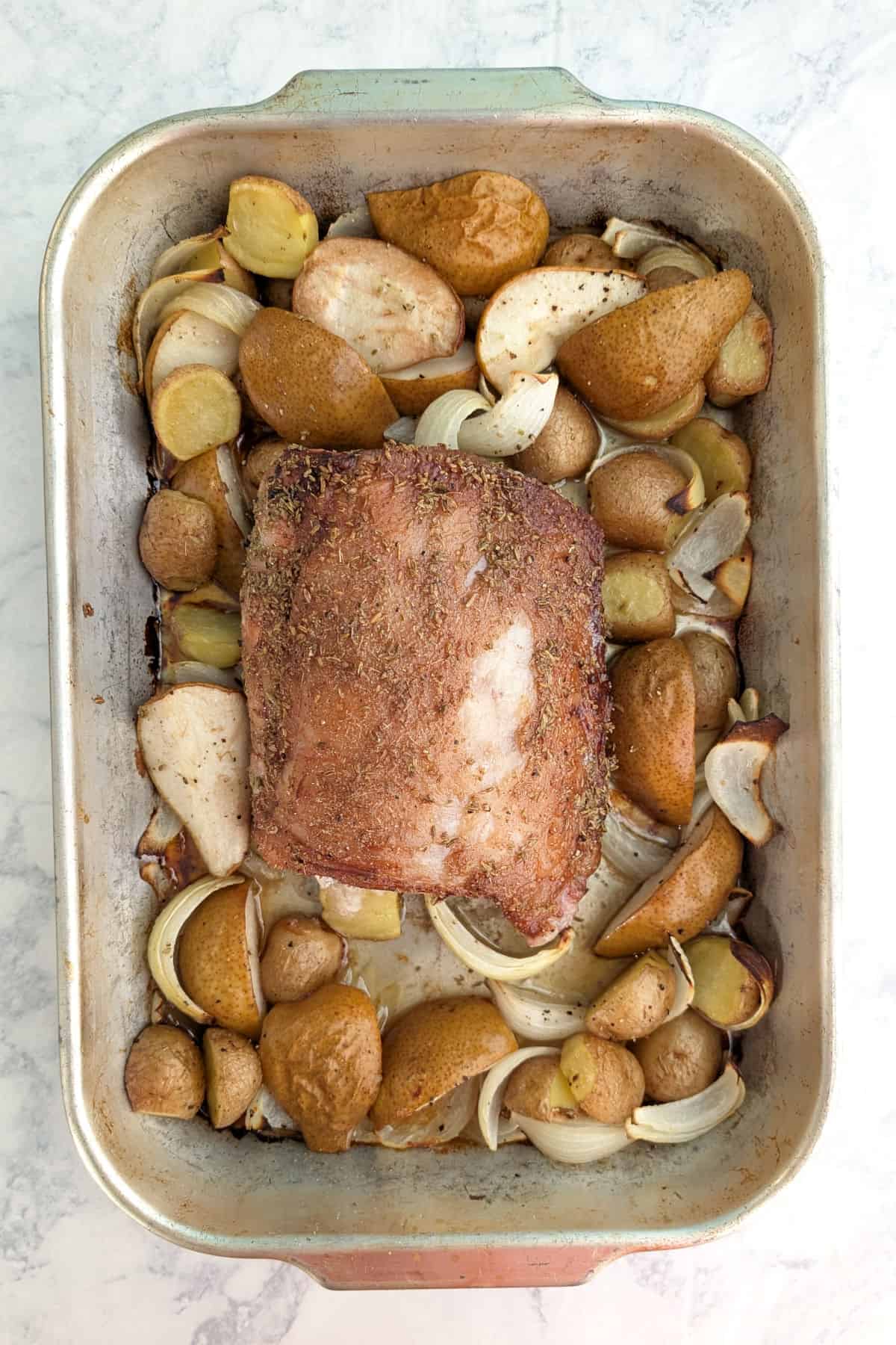 Roasted fennel-coated pork with pears, potatoes, and onions, in a roasting pan.