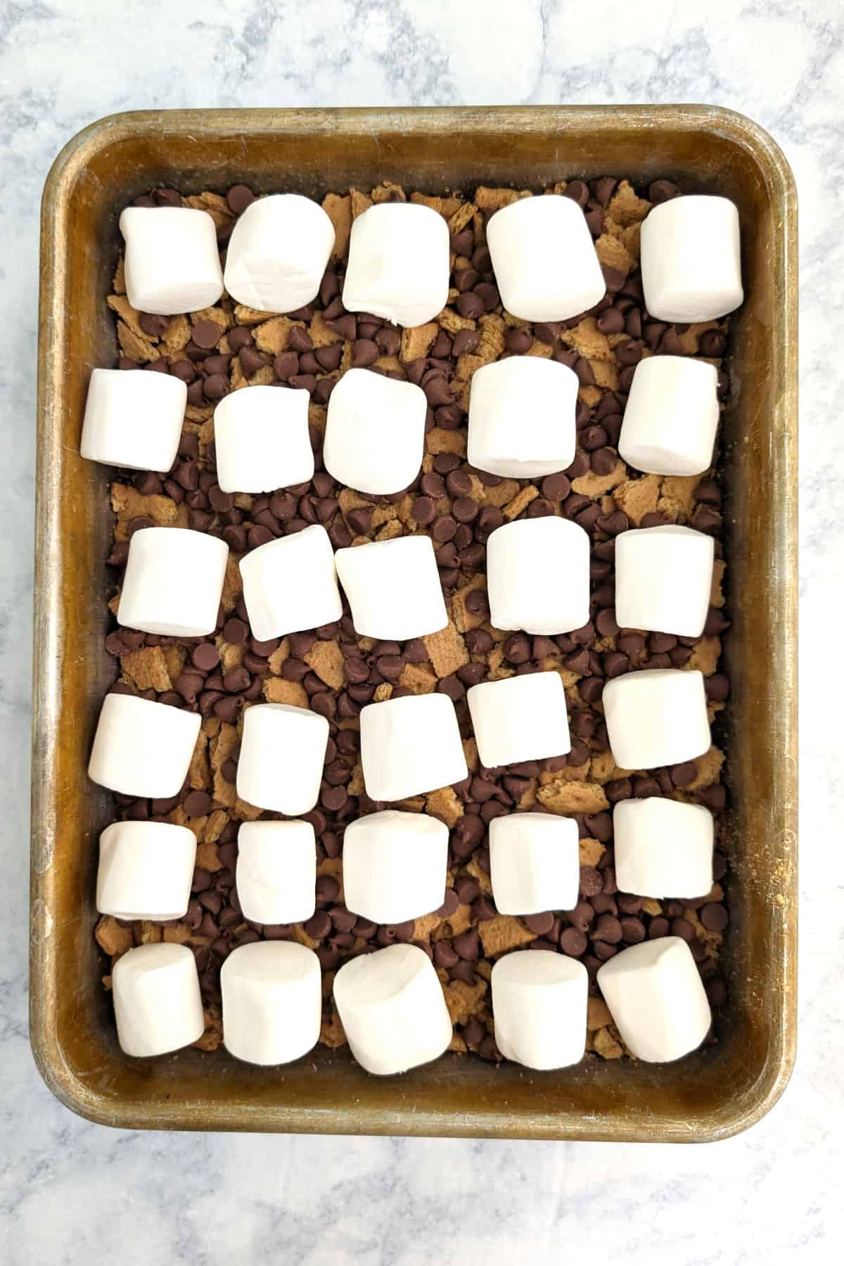 Brownie batter, in a pan, topped with crushed graham crackers, milk chocolate chips, and marshmallows.