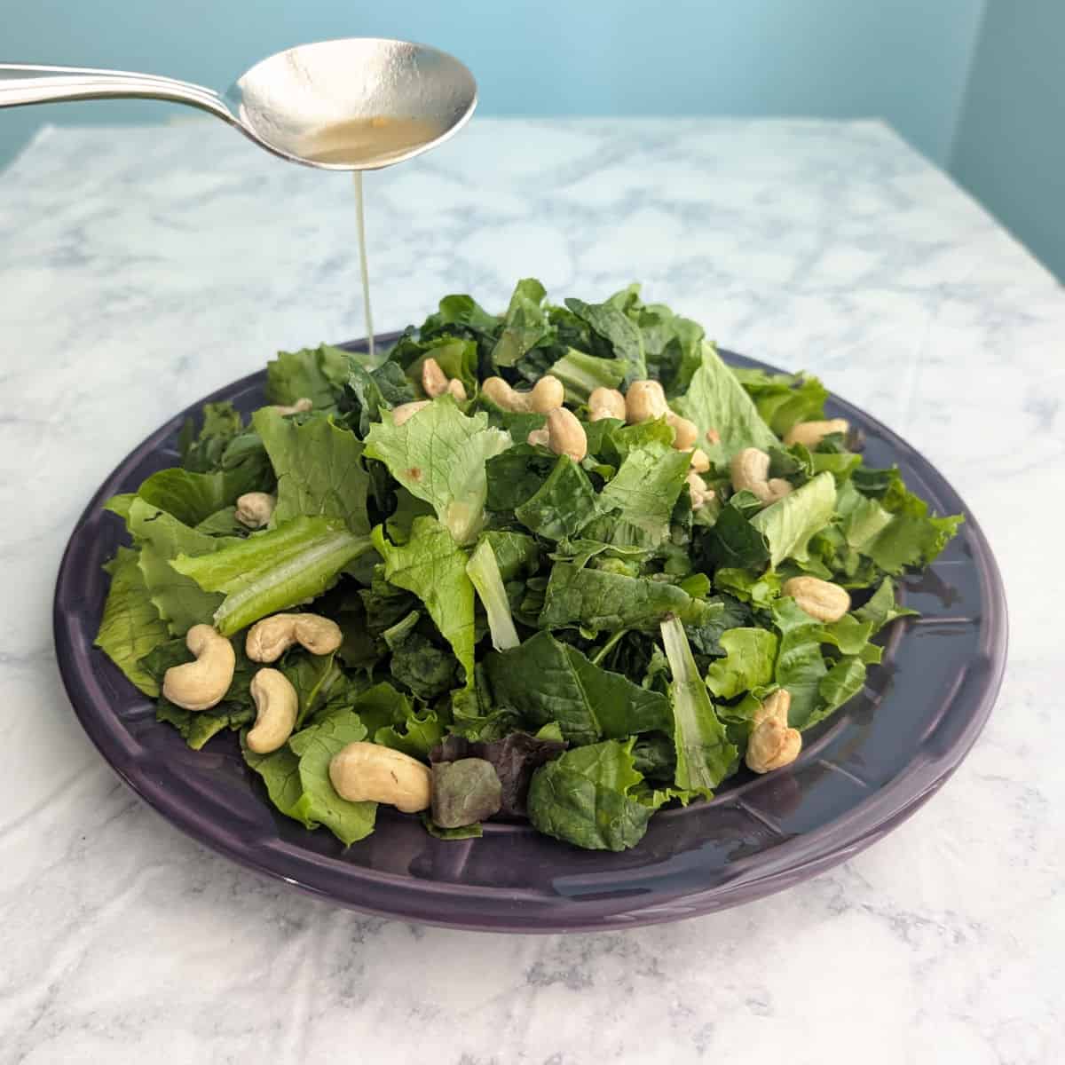 Salad dressing drizzles from a spoon onto salad greens and cashews on a purple plate.