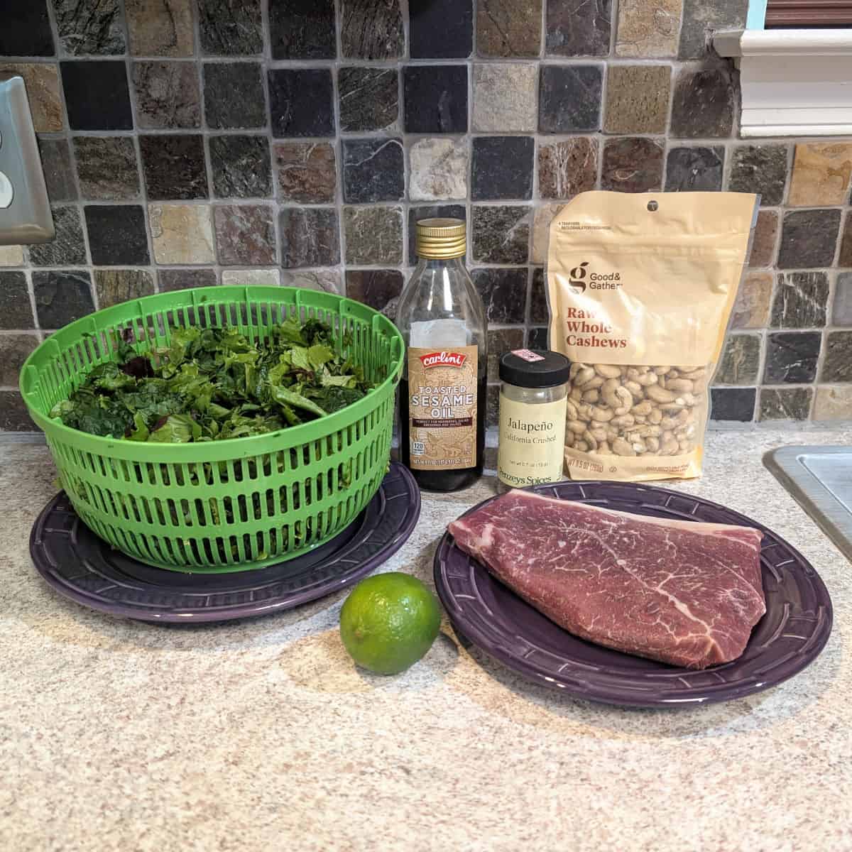 Salad mix in a salad spinner, sesame oil, jalapeno flakes, cashews, a lime, and an uncooked steak.