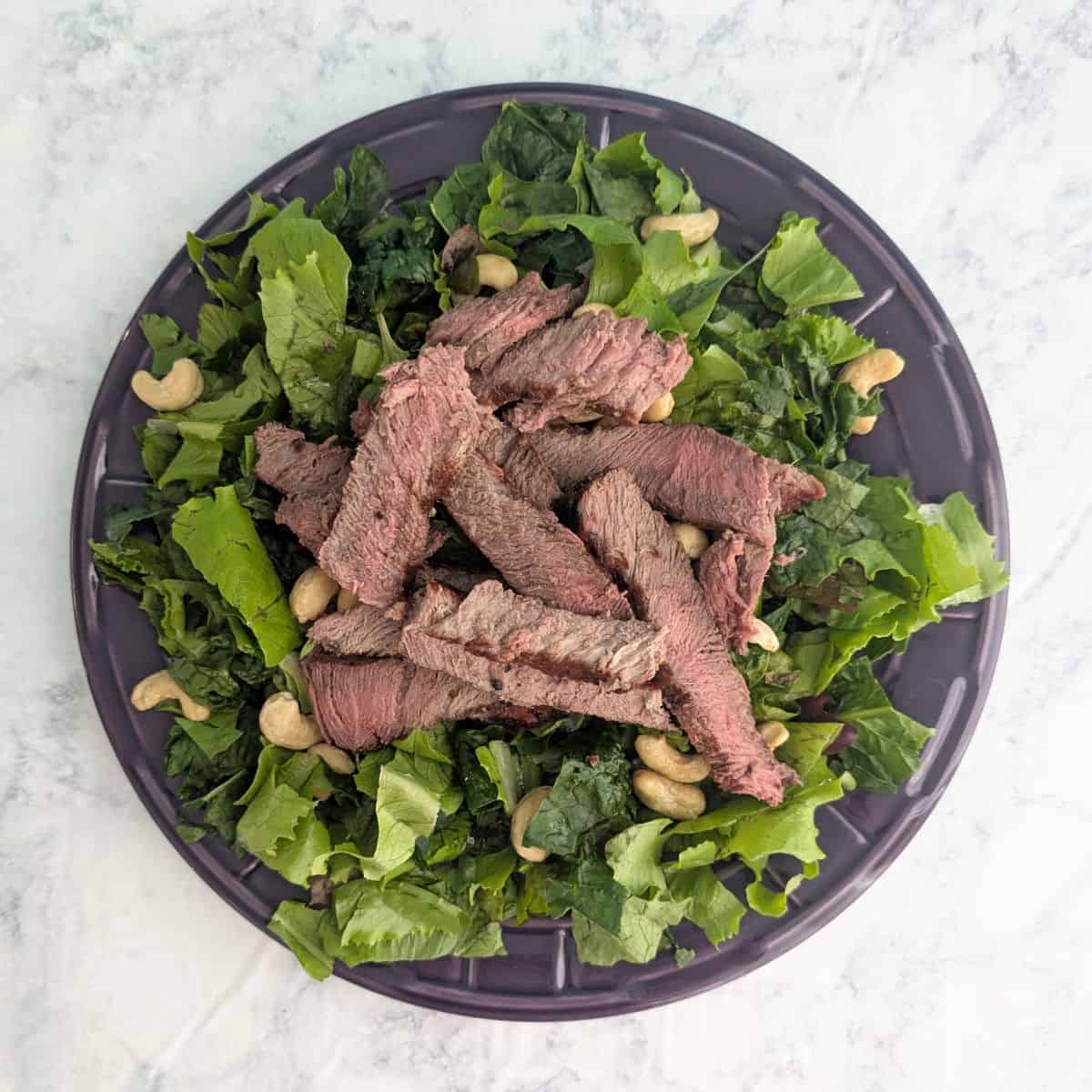 Grilled, sliced steak and cashews on salad greens on a purple plate.
