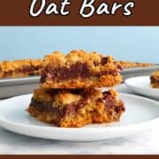 A stack of 2 chocolate oat bars on a plate, in front of a pan of oat bars. Text overlay for Pinterest reads "Chocolate Oat Bars. Rich and Decadent!"