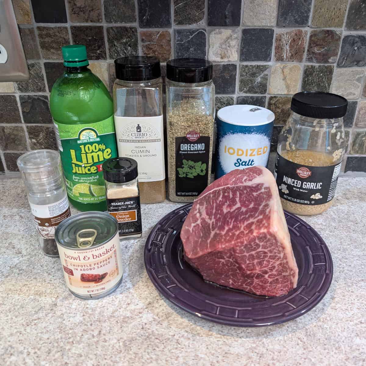 Ingredients for barbacoa: beef roast, black pepper, lime juice, onion powder, a can of chipotle peppers in adobo sauce, ground cumin, dried oregano, salt, a jar of minced garlic.