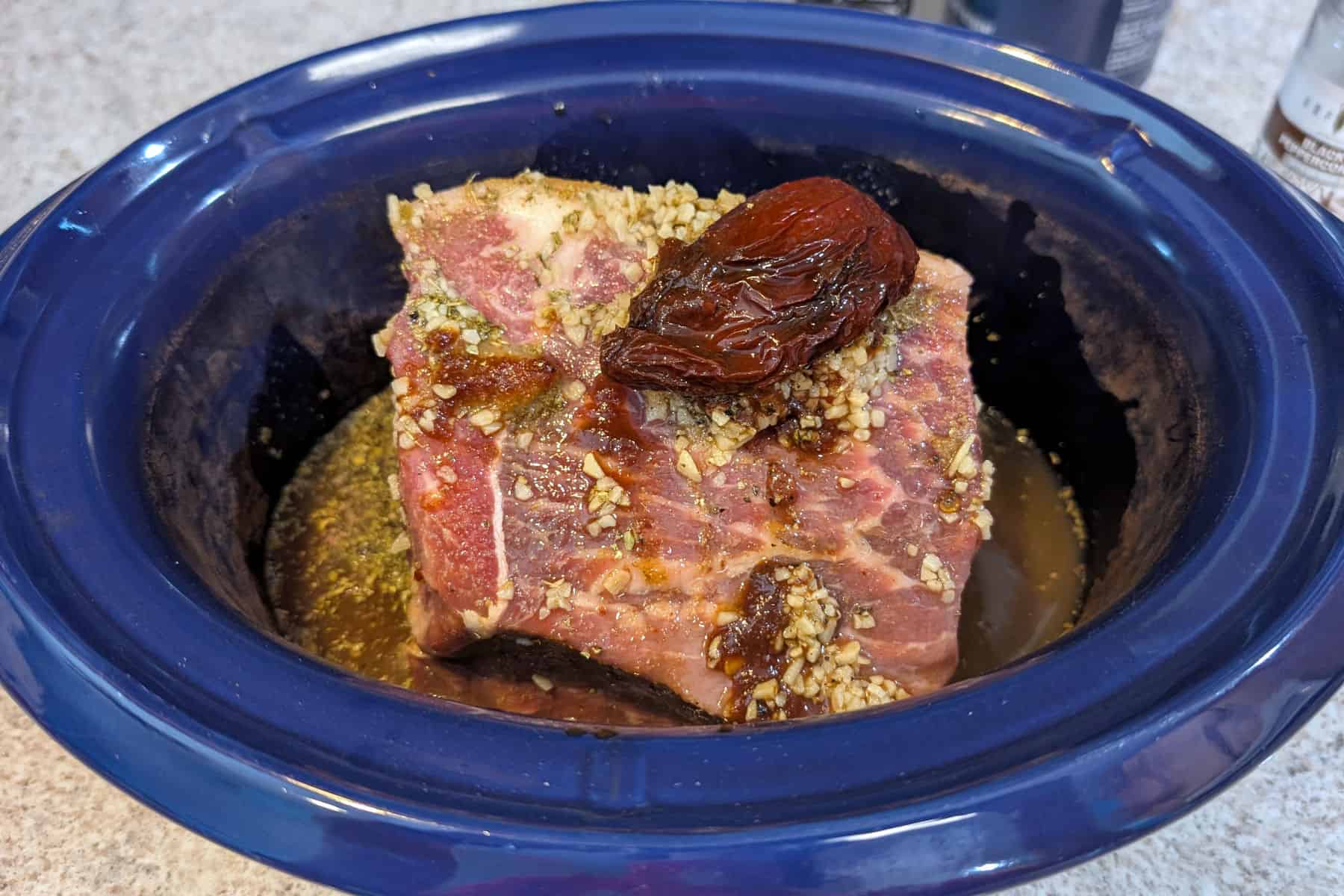 An uncooked beef roast with garlic and a chipotle on top, in the crock of a 3.5 quart slow cooker, with liquid at the bottom.