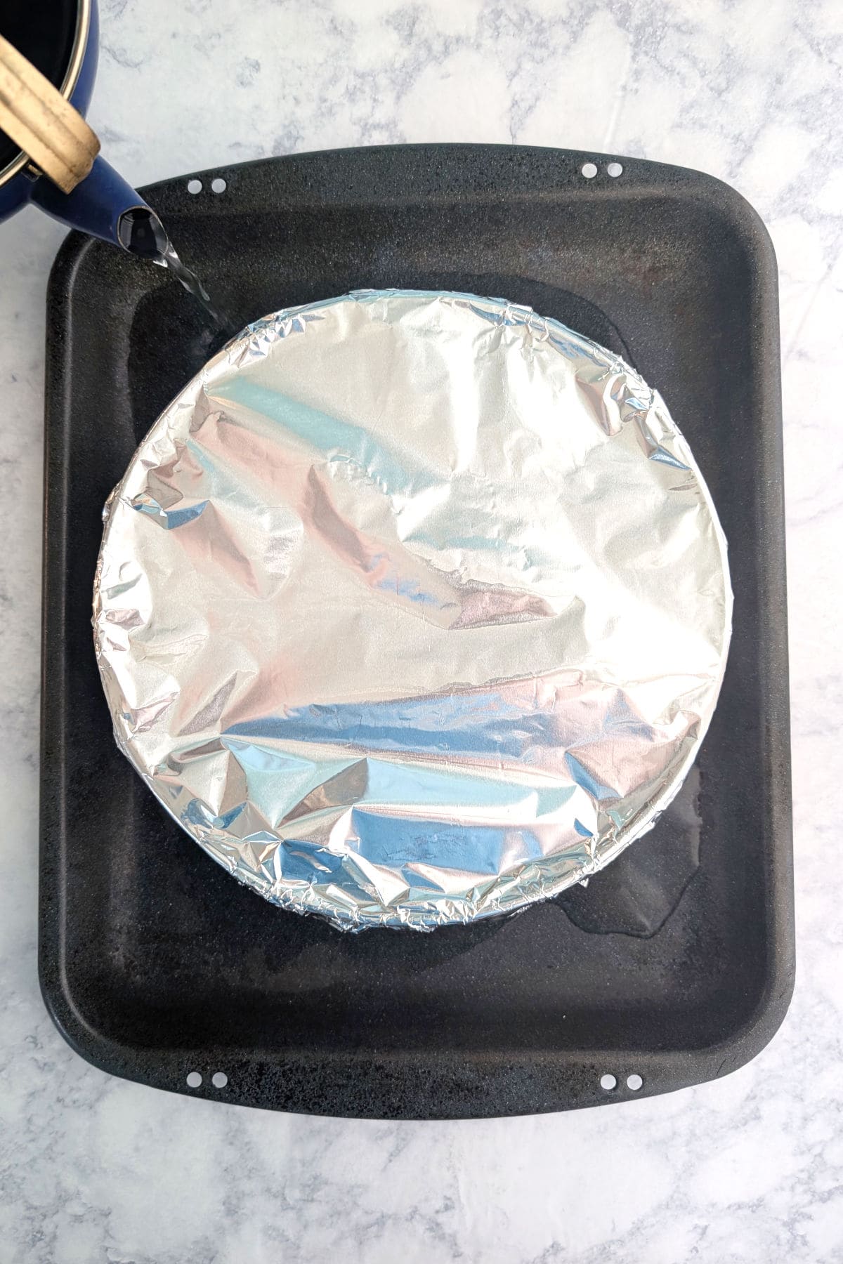 A Springform pan covered with aluminum foil, in a larger roasting pan. Water is poured from a kettle into the roasting pan to make a water bath.