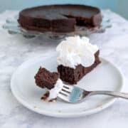 A bite of chocolate truffle cake on a fork, next to a slice of cake with whipped cream. The remaining cake is on a plate in the background.