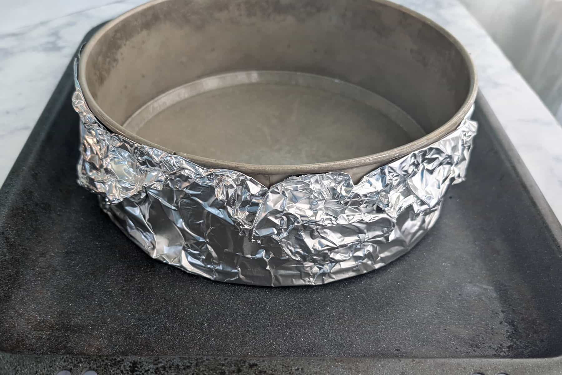 The bottom of a Springform pan is wrapped in aluminum foil. It sits in a larger roasting pan that will later have water to create a water bath.