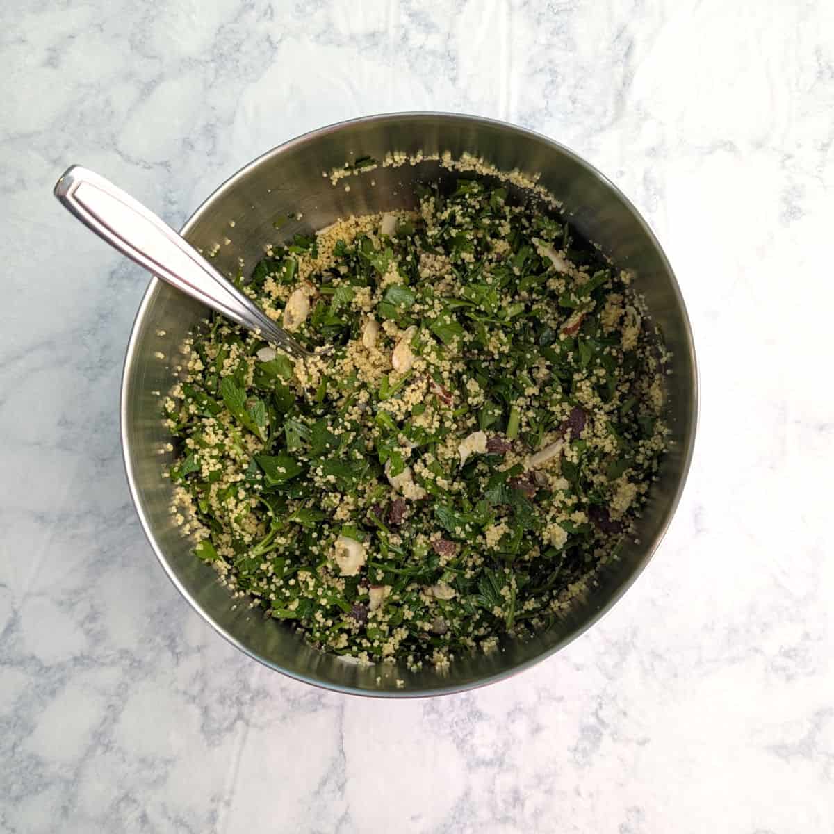 Couscous and parsley mixed with almonds, raisins, and capers in a mixing bowl.