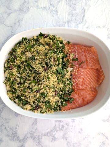 Couscous with parsley partially covers raw salmon in a baking dish.