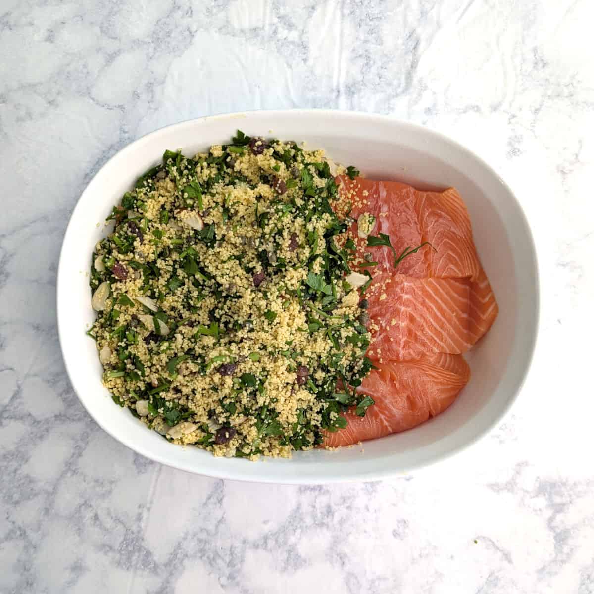 Couscous with parsley partially covers raw salmon in a baking dish.