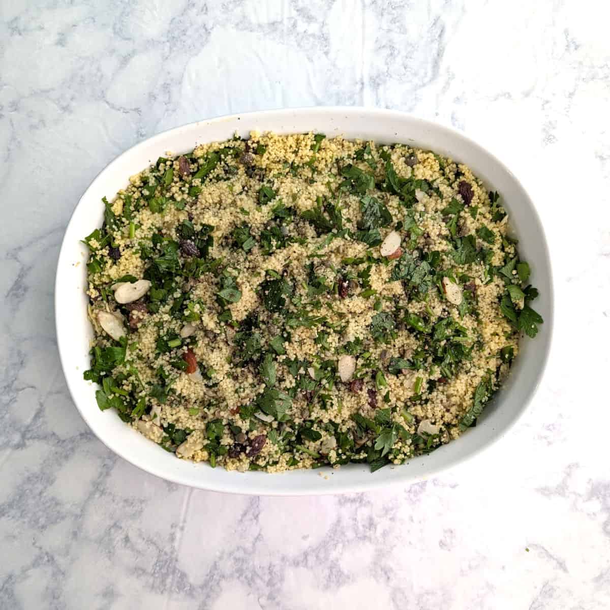 Couscous and parsley with almonds, raisins, and capers, covering salmon in a white baking dish.