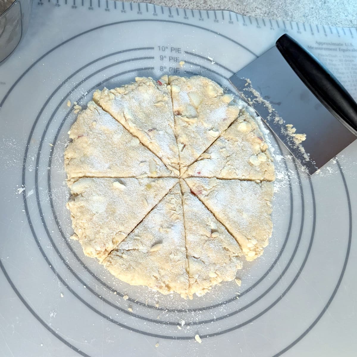 Dough for Apple and Pear Scones, patted into a 9 inch circle, sprinkled with sugar, and cut into 8 wedges. A bench scraper rests next to the dough on a pastry mat.