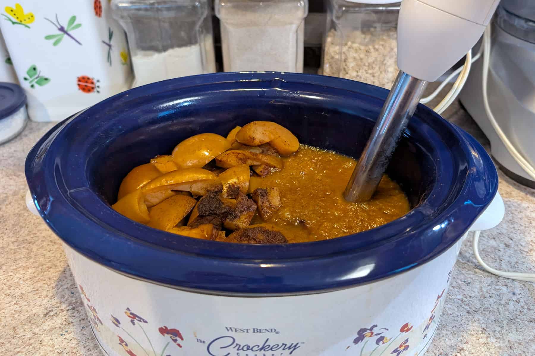 Cooked apples and spices in a slow cooker. An immersion blender rests in it, and has pureed apples in the right side of the crock.