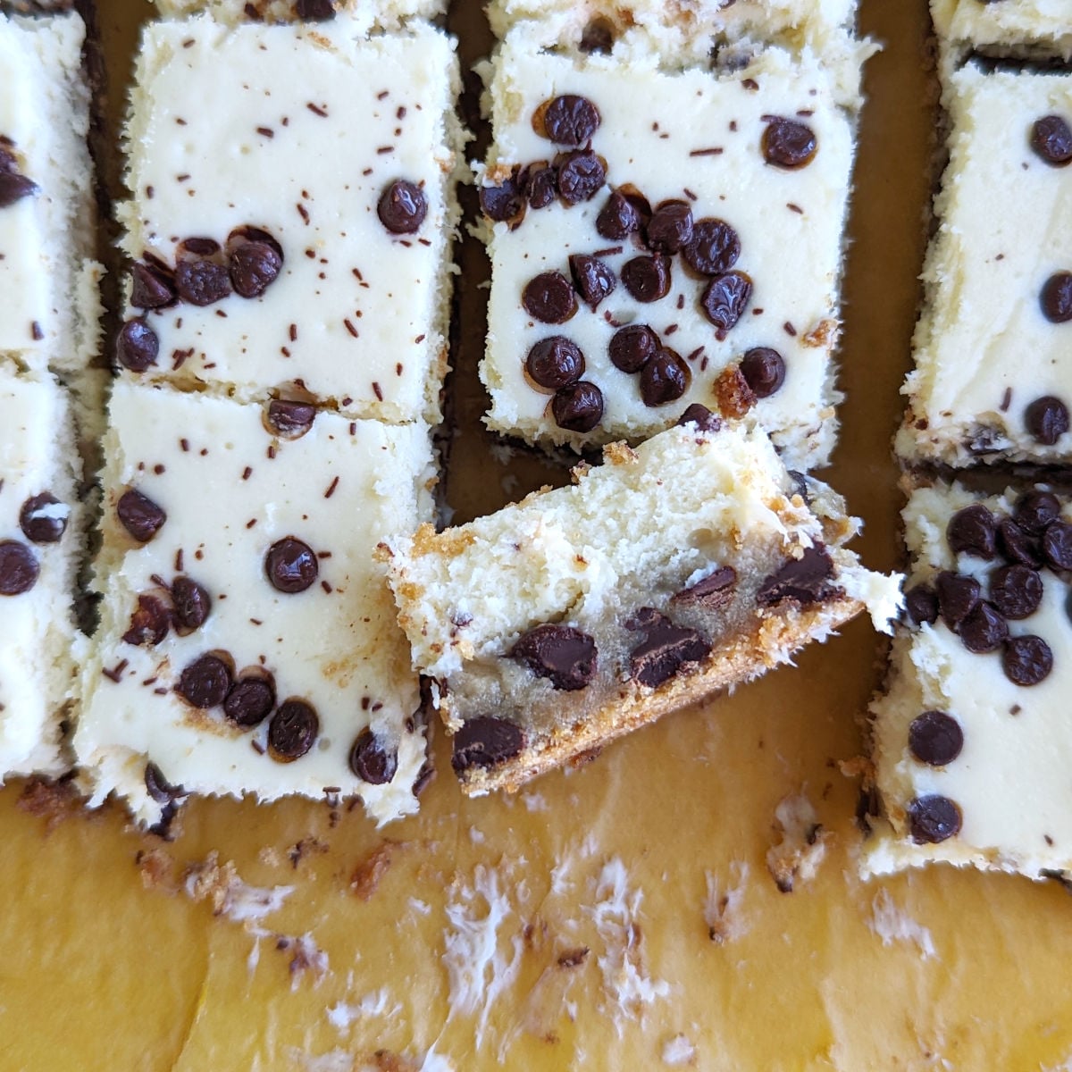 Chocolate chip cookie cheesecake bars, cut into squares. They are viewed from above, and one is propped on its side to show the chocolate chip cookie layer and the cheesecake layer topping it.