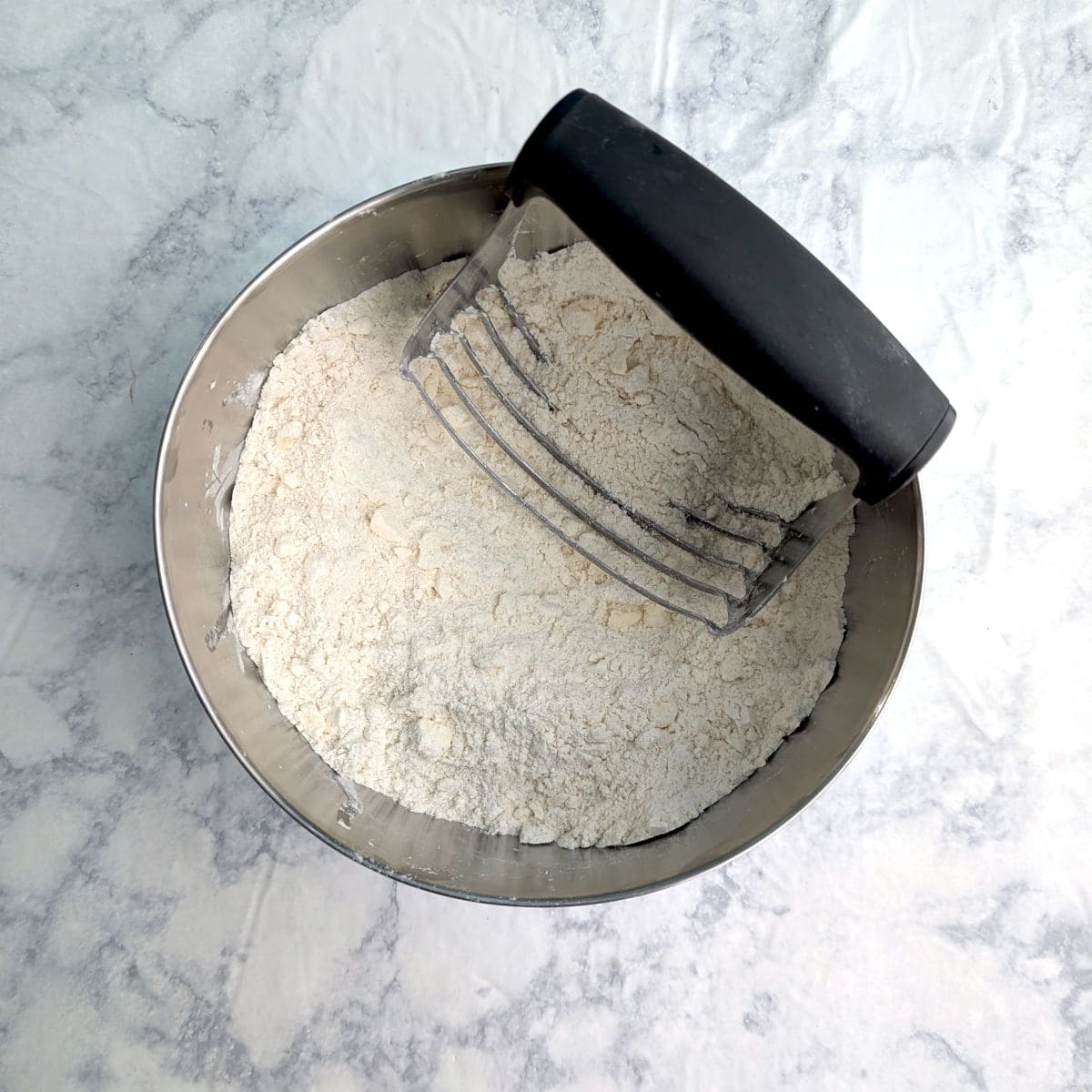 In a bowl, butter has been cut in a flour mixture into small, crumb-sized pieces. A pastry cutter rests in the bowl.