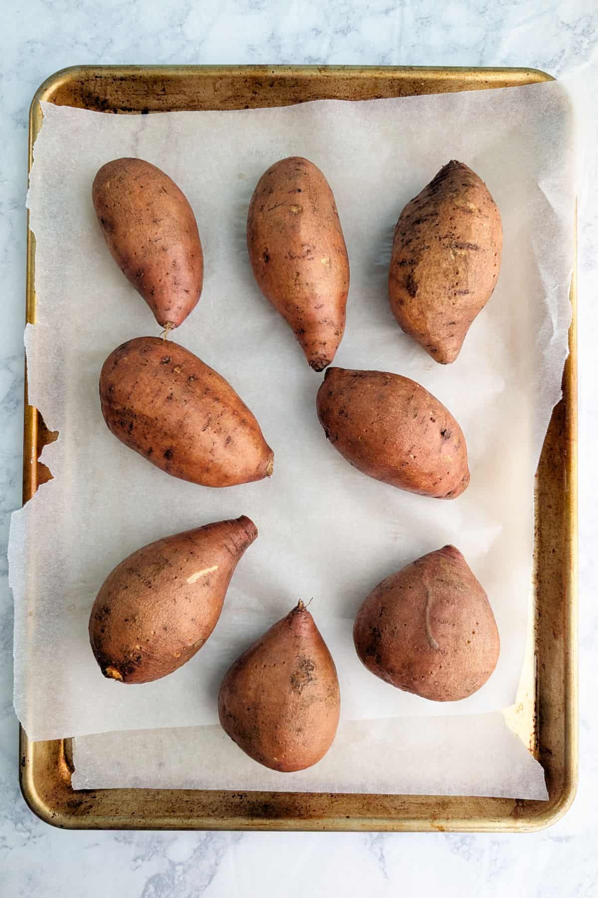 Raw whole sweet potatoes, pricked by a fork, on a baking sheet lined with parchment paper.