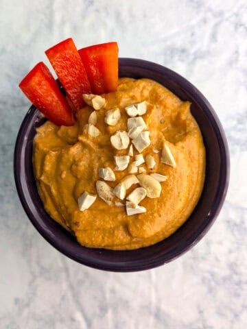 Smoky Cashew Dip, garnished with chopped cashews and red bell pepper strips, in a small purple ramekin.