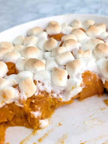 Lightly browned marshmallows on top of soft sweet potatoes in a casserole dish. Half of the sweet potatoes have been served, leaving the other half behind.