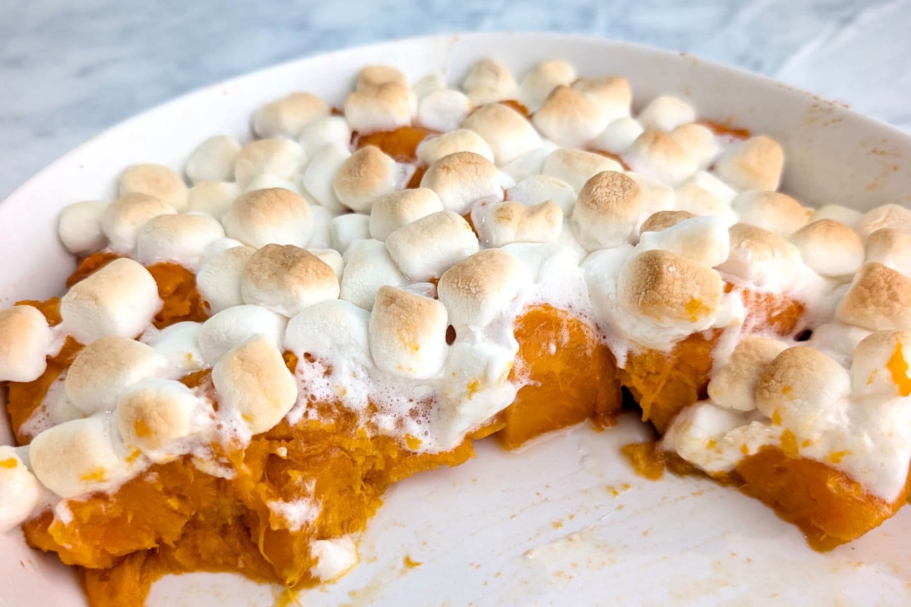 Lightly browned marshmallows on top of soft sweet potatoes in a casserole dish. Half of the sweet potatoes have been served, leaving the other half behind.