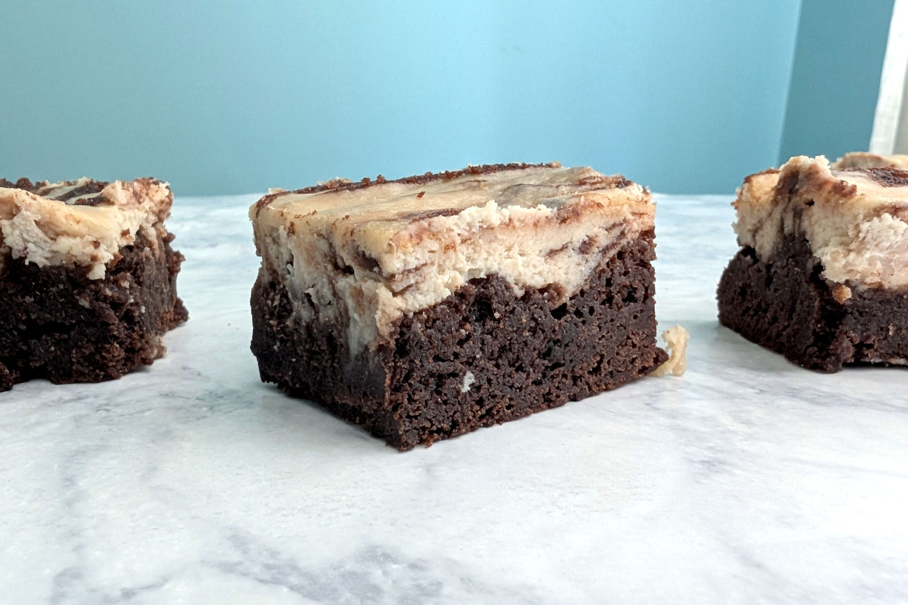 A cheesecake brownie with cheesecake brownies on each side, seen from a side angle to show distinct layers of cheesecake on top of brownie.