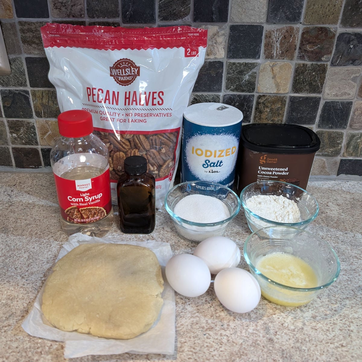 Ingredients for pecan fudge pie: corn syrup, pecans, vanilla, salt, sugar, cocoa powder, flour, eggs, melted butter, and pie crust dough.