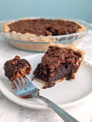 A fork with a bite of Pecan Fudge Pie rests on a plate with the slice. The remaining pie is in the background.