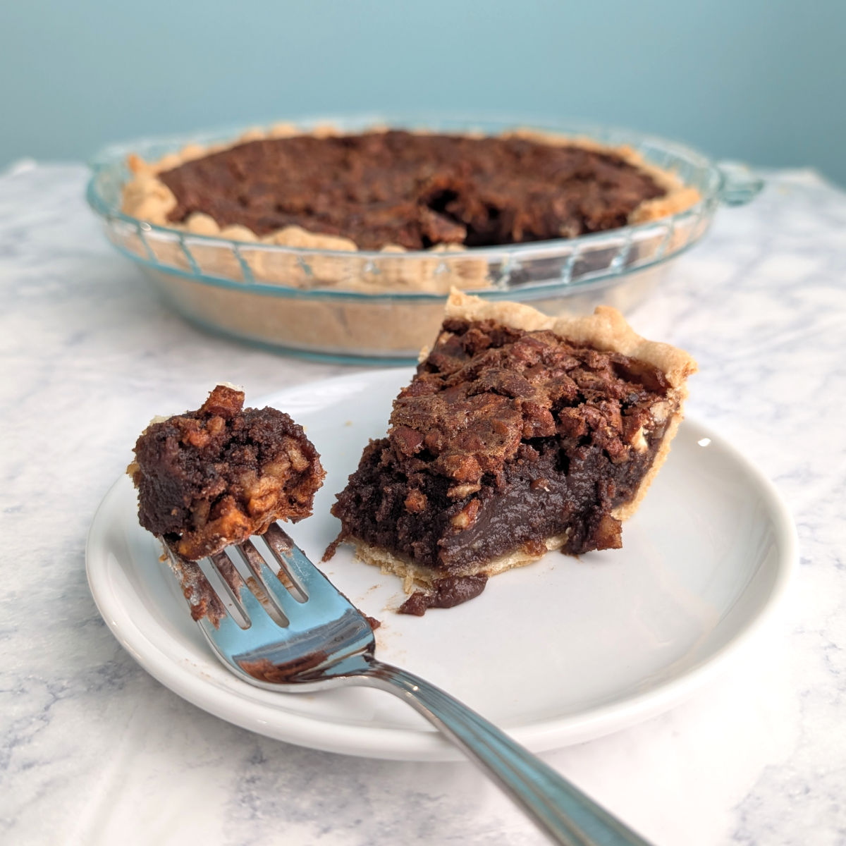 A fork with a bite of Pecan Fudge Pie rests on a plate with the slice. The remaining pie is in the background.