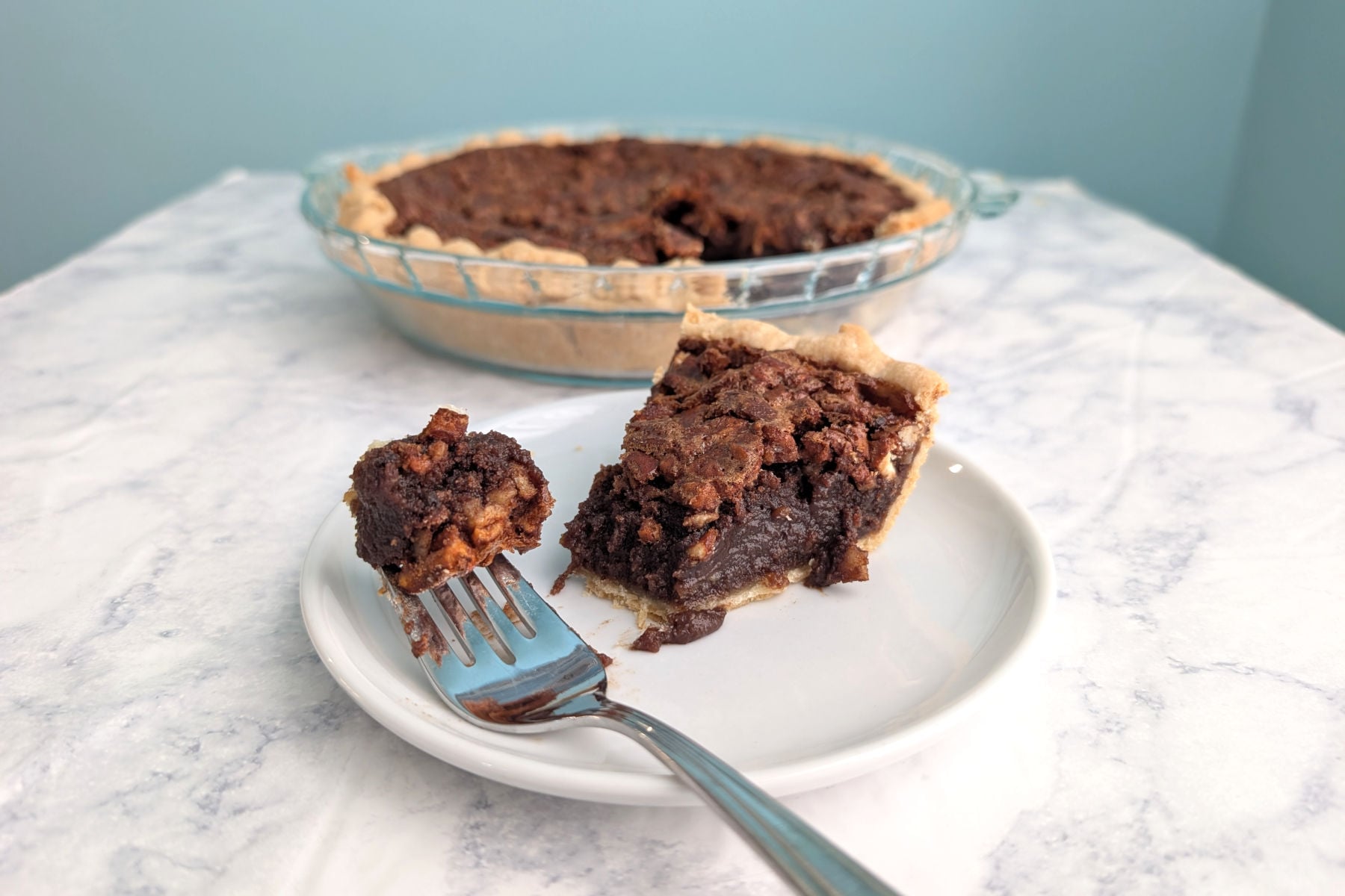 A fork with a bite of Pecan Fudge Pie rests on a plate with the slice. The remaining pie is in the background.
