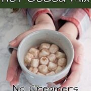 Hands hold a mug of hot cocoa topped with miniature marshmallows. Text overlay for Pinterest reads "Homemade Hot Cocoa Mix. No Creamers. Just Cocoa. Easy As Cookies"